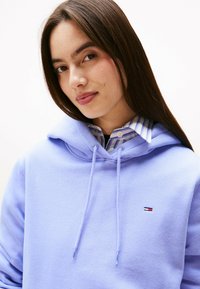 Młoda kobieta z długimi brązowymi włosami, nosząca jasnofioletowy hoodie na niebiesko-białą koszulę z kołnierzykiem, delikatnie patrzy w kamerę.