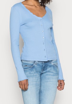 Femme portant un cardigan ajusté bleu clair côtelé à boutons avec des bords festonnés et un jean bleu clair délavé.