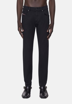 Man draagt zwarte slim-fit jeans en zwarte enkellaarzen met gespen, staand tegen een effen achtergrond.