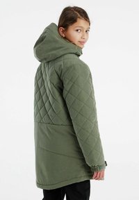 Giacca trapuntata verde con cappuccio, caratterizzata da un motivo tratteggiato e polsini elasticizzati. Il materiale sembra caldo e resistente, adatto per il freddo.