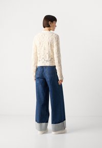Cremfarbener gehäkelter Pullover mit taillierter Passform, kombiniert mit weit geschnittenen blauen Jeans aus Denim mit einem kontrastierenden Saum. Einfache weiße Sneakers.