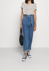 Blusa crema con macchie nere, jeans in denim blu a vita alta con cintura in vita, e sneakers nere alte. Borsa intrecciata nera inclusa.
