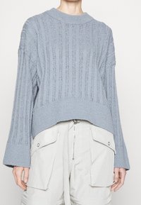 Maglione lavorato a maglia di colore azzurro chiaro con coste verticali, maniche ampie e orlo corto, abbinato a pantaloni cargo chiari con tasche.