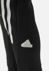 Pantaloni sportivi neri con tre righe bianche lungo i lati e un logo Adidas grigio sulla coscia sinistra. Tessuto liscio e elasticizzato.