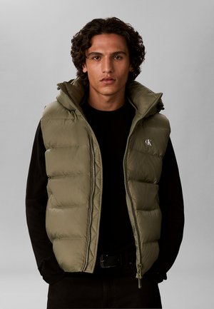 Junger Mann mit lockigem Haar, der eine olivgrüne gesteppte Weste über einem schwarzen Langarmshirt trägt, steht vor einem einfarbigen grauen Hintergrund.