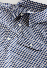 Chemise à col boutonné avec un motif à carreaux bleu, beige et blanc, comprenant une poche avant avec rabat boutonné.