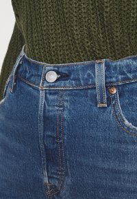 Modré denimové vysoké džínsy s měděným prošíváním, stříbrným zapínáním a mírným zesvětlením na stehnech, zkombinované s tmavě zeleným pleteným topem.