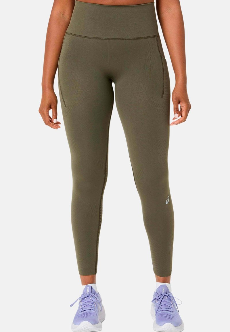 Close-up of person wearing olive green high-waist leggings with side pockets and purple running shoes on a white background.