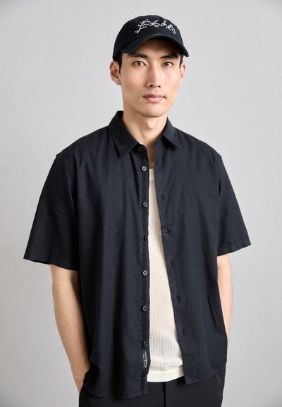 Čierna krátka mäkká košeľa vyrobená z ľahkého materiálu, so štrukturálnym dizajnom, so zapínaním na gombíky a otvoreným golierom. Pod ňou je biely sieťovaný top.