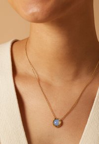 Gouden ketting met een ronde, blauwe stenen hanger, gezet in een cirkelvormig gouden frame, gedragen tegen een lichte huidtoon.
