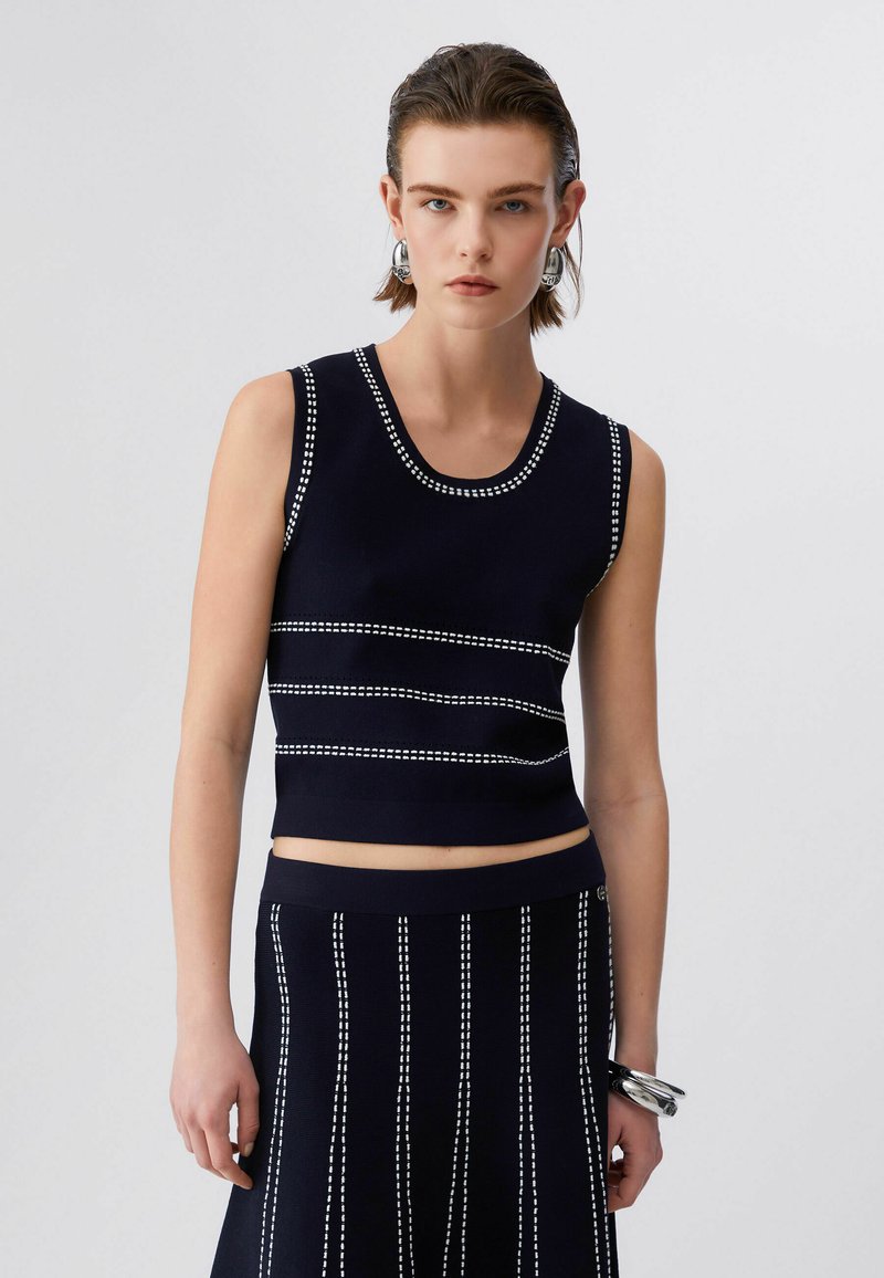 Femme portant un haut court noir sans manches et une jupe assortie avec des rayures blanches cousues, accessoirisée avec des boucles d'oreilles et des bracelets argentés.