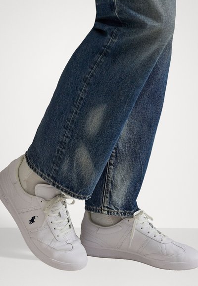 Balti sportiniai batai su lygia odos apdaila, su raišteliais ir šoninėmis juostelėmis, dėvimi su tamsiai mėlynomis džinsinėmis kelnėmis.