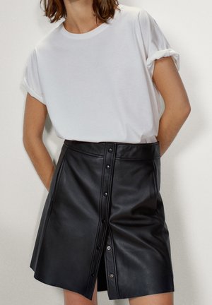Person wearing a short-sleeve white t-shirt tucked into a black button-front leather skirt against a plain background.