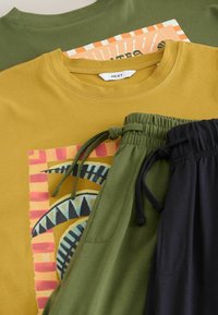 T-shirt graphique moutarde avec motif géométrique, pantalon à cordon olive vert et noir, et t-shirt olive vert avec un design orange et blanc en couche en dessous.