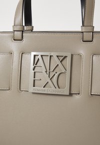 Borsa a tracolla sintetica beige con finitura testurizzata, caratterizzata da una prominente placca logo grigia e design strutturato. Due manici robusti attaccati.