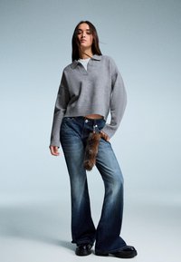 Grauer, kürzer geschnittener Pullover mit Kragen, kombiniert mit blauen Schlagjeans, die einen braunen Fell-Schlüsselanhänger haben. Schwarze Plateauschuhe runden das Outfit ab.