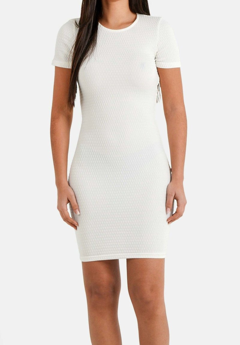 Femme portant une robe blanche texturée ajustée, à manches courtes, longueur genoux, debout devant un fond clair uni.