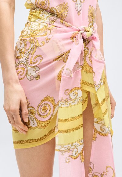 Τα χέρια του μοντέλου ακουμπούν σε μια ροζ και κίτρινη φούστα με φουλάρι, δεμένη, που φέρει περίτεχνα χρυσά λουλουδένια και σκαλιστά μοτίβα, αποκαλύπτοντας μια σκίσιμο στο πόδι.