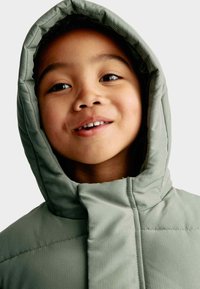 Polstret grøn hoodie med en struktureret overflade, der har en høj krave og en tætsiddende pasform omkring ansigtet, som fremhæver et barns glade udtryk.