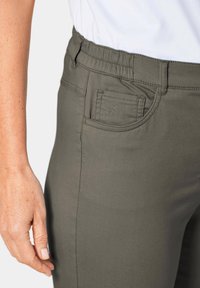 Elastische taillebroek in een gedempte groene kleur, gemaakt van zacht materiaal. Beschikt over een achterzak met ton-sur-ton stiksels en een gladde textuur.