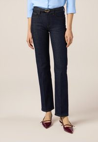 Jeans de mezclilla azul oscuro con diseño de pierna recta, que cuentan con una hebilla de cinturón circular dorada y costuras de contraste. Llevados con tacones de charol burdeos.