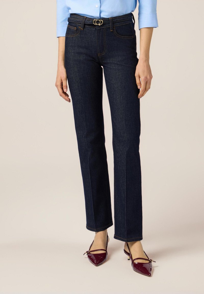 Jeans de mezclilla azul oscuro con diseño de pierna recta, que cuentan con una hebilla de cinturón circular dorada y costuras de contraste. Llevados con tacones de charol burdeos.