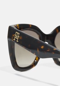 Gafas de sol de carey con un marco geométrico grueso, lentes con degradado y un acento dorado con el logo en la patilla. Textura mate, diseño audaz.
