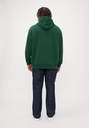 Persoon met gevlochten haar, gekleed in een donkergroene hoodie, donkerblauwe jeans en witte schoenen, met de rug naar voren tegen een effen lichte achtergrond.
