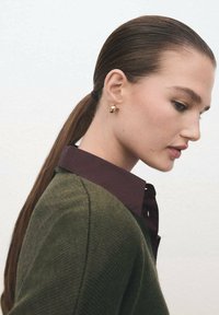 Los pendientes de tuerca dorados complementan un suéter de punto verde oscuro con una camisa de cuello burdeos. El cabello liso está peinado en una coleta baja.