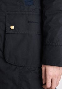 Detail ruky vedle kapsy černé bundy Barbour se zlatým patentním knoflíkem a vyšitým názvem značky.