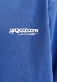Sudadera de cuello redondo de algodón azul con un gráfico blanco que dice "project core" junto con "JACK & JONES © 1990 CREATIVE."