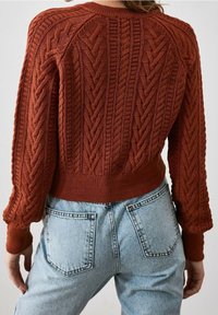 Rust-colored knit sweater with textured cable patterns, ribbed cuffs and hem, paired with light blue high-waisted denim jeans.