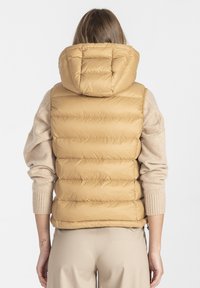 Beżowa, puchowa kamizelka z kapturem, z pikowanym wzorem i gładką fakturą. Nosi się ją na beżowym swetrze z prążkowanymi mankietami.