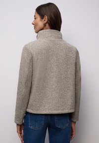 Grau strukturiertes Fischgrätmuster-Jackett mit hohem Kragen, figurbetontem Design und sichtbaren Nähten, kombiniert mit blauen Jeans.