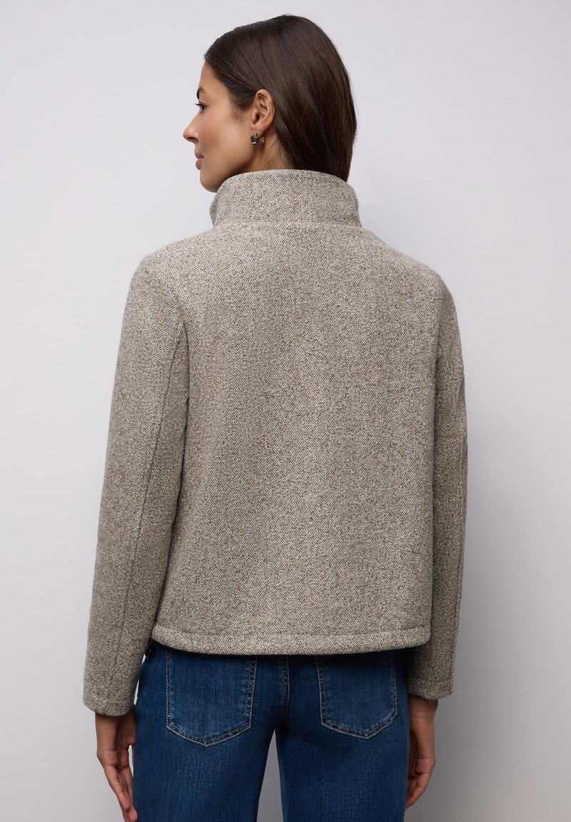 Szara teksturowana marynarka w jodełkę z wysokim kołnierzem, dopasowanym krojem i widocznymi detalami szwów, zestawiona z niebieskimi dżinsami.