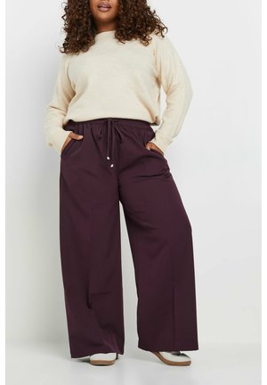 Beige Pullover mit langen Ärmeln, kombiniert mit weit geschnittenen, dunkelvioletten Hosen mit elastischem Bund und seitlichen Taschen.