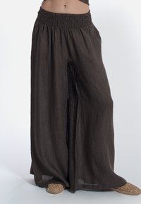 Pantaloni larghi in lino marrone con cintura smock; tessuto leggero con un design fluido, abbinati a scarpe slip-on intrecciate.