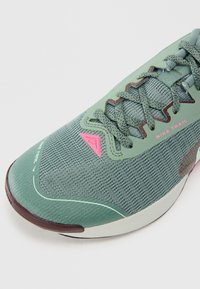 Chaussure de trail Nike de couleur vert clair avec un upper en mesh texturé, des accents roses et une semelle en caoutchouc marron foncé. Comprend des lacets et des détails du logo.