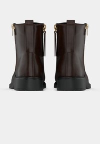 Botas de cano baixo em pele castanha com solas de borracha pretas, apresentando detalhes em metal dourado e puxadores de fecho éclair decorativos na parte de trás.