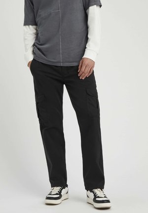 Pantalons cargo noirs avec plusieurs poches latérales, associés à un t-shirt gris à manches courtes sur une chemise blanche à manches longues, et des baskets noir et blanc.