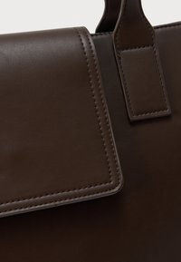 Καφέ σκούρα δερμάτινη τσάντα χειρός με λεπτομερείς ραφές στη φλάντζα και στην πρόσδεση της λαβής, που αναδεικνύουν την απαλή υφή και την τέχνη κατασκευής.