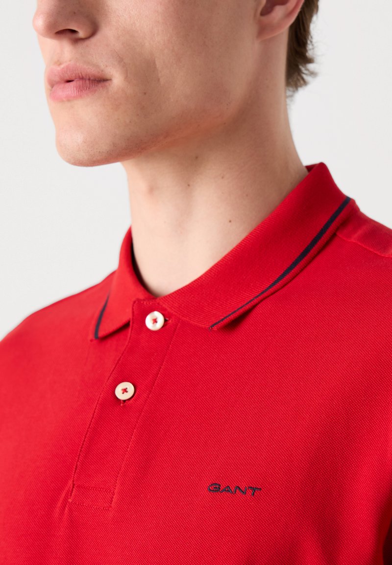 Polo rouge avec une finition texturée, deux boutons blancs et un liseré de col bleu marine. Présente un petit logo bleu sur la poitrine.