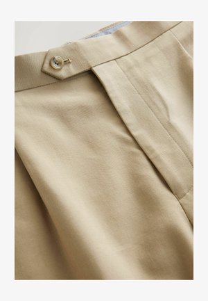 Pantalons beige avec une taille structurée, fermeture à un bouton et plis devant. Le tissu a une texture lisse et un motif diagonal subtil.