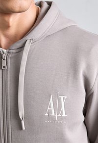 Felpa con cappuccio grigia con zip, realizzata in tessuto liscio, dotata di cappuccio, zip frontale e logo ricamato "A|X". Texture discreta con dettagli minimi.