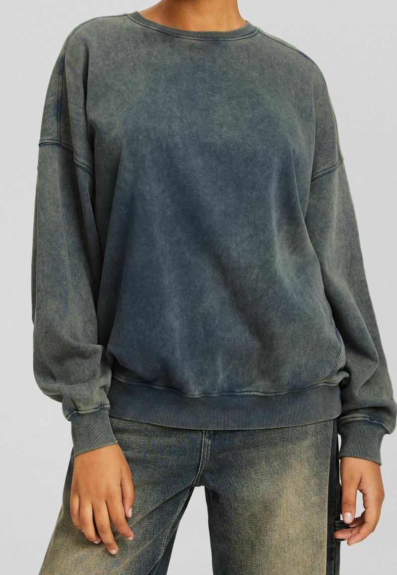 Sweatshirt en tissu bleu foncé délavé avec une coupe décontractée, un col rond et des poignets côtelés. Présente une finition douce et texturée.