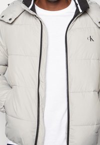 Giacca puffer grigia chiara con zip frontale, accenti neri, cappuccio staccato e logo ricamato sul lato sinistro del petto. Superficie testurizzata.