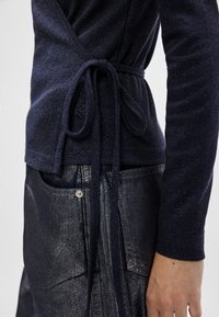 Granatowy sweter w formie owijanej z teksturowanym splotem, z wiązaniem w pasie. Noszony nad ciemną, błyszczącą spódnicą dżinsową z przednią kieszenią.