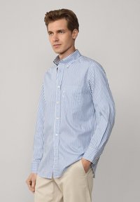 Camisa de cuadros en azul claro y blanco, con cuello, mangas largas y un bolsillo en el pecho. La tela es de algodón suave.
