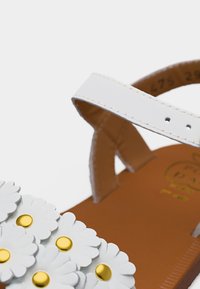 Sandale en cuir blanc avec bride de cheville et décorations en forme de marguerite avec des centres jaunes sur une semelle marron.