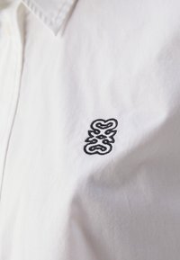 Chemise blanche avec un col classique, ornée d'un logo noir brodé sur le côté gauche de la poitrine. Texture de tissu lisse avec un design épuré.